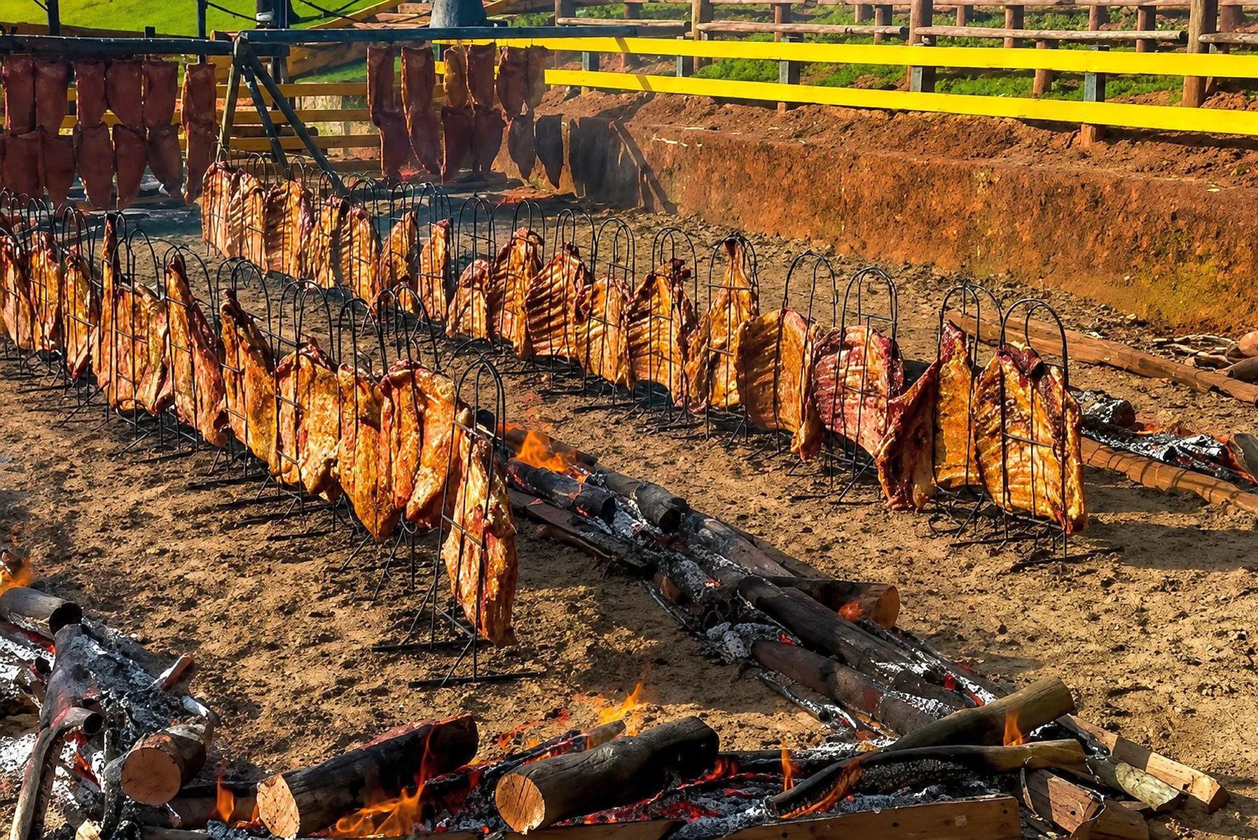 imagem mostra dezenas de costelas alinhada se presas a espetos, no chão, com lenhas queimando ao redor
