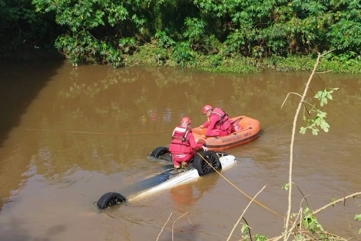 Imagem mostra um carro submerso em um rio com um barco dos Bombeiros ao lado.