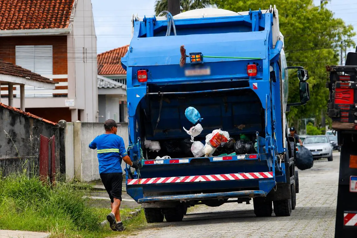 Imagem mostra um caminhão de lixo azul com um coletor correndo atrás.