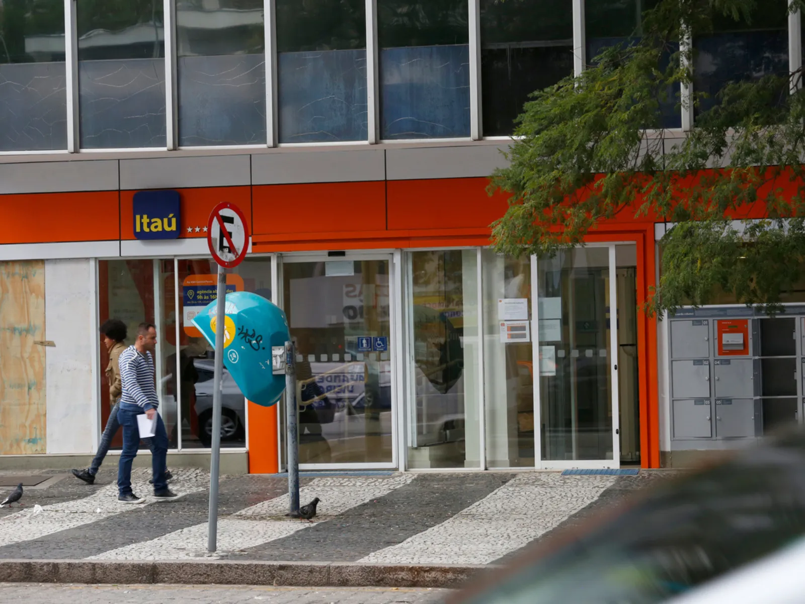 Imagem mostra um prédio em Curitiba com a fachada vermelha, com uma pessoa e um telefone público na frente. Tem uma placa de proibido estacionar e galhos de árvore.