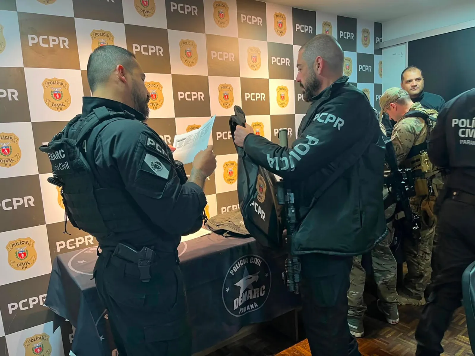 Fotografia mostra policiais civis durante uma operação. Em primeiro plano, dois agentes vestidos de preto conversam diante de uma mesa, enquanto um deles segura e consulta um documento. Ao fundo, outros policiais, alguns usando uniformes camuflados e equipamentos táticos, aparecem próximos à parede. O ambiente tem um painel quadriculado com os logotipos da Polícia Civil e, na mesa, há uma identificação da Denarc Paraná.