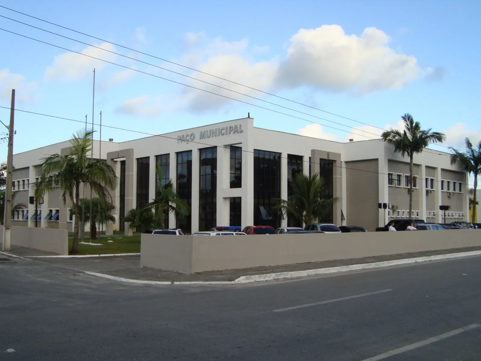 Fotografia da sede da Prefeitura de Matinhos. A foto mostra a fachada de um prédio branco de dois andares, identificado na parte superior como “Paço Municipal”. O edifício tem grandes janelas de vidro escuro e detalhes em tons claros na fachada. Em frente, há um jardim com palmeiras, uma área cercada por um muro baixo e vários carros estacionados. A construção fica às margens de uma avenida asfaltada.
