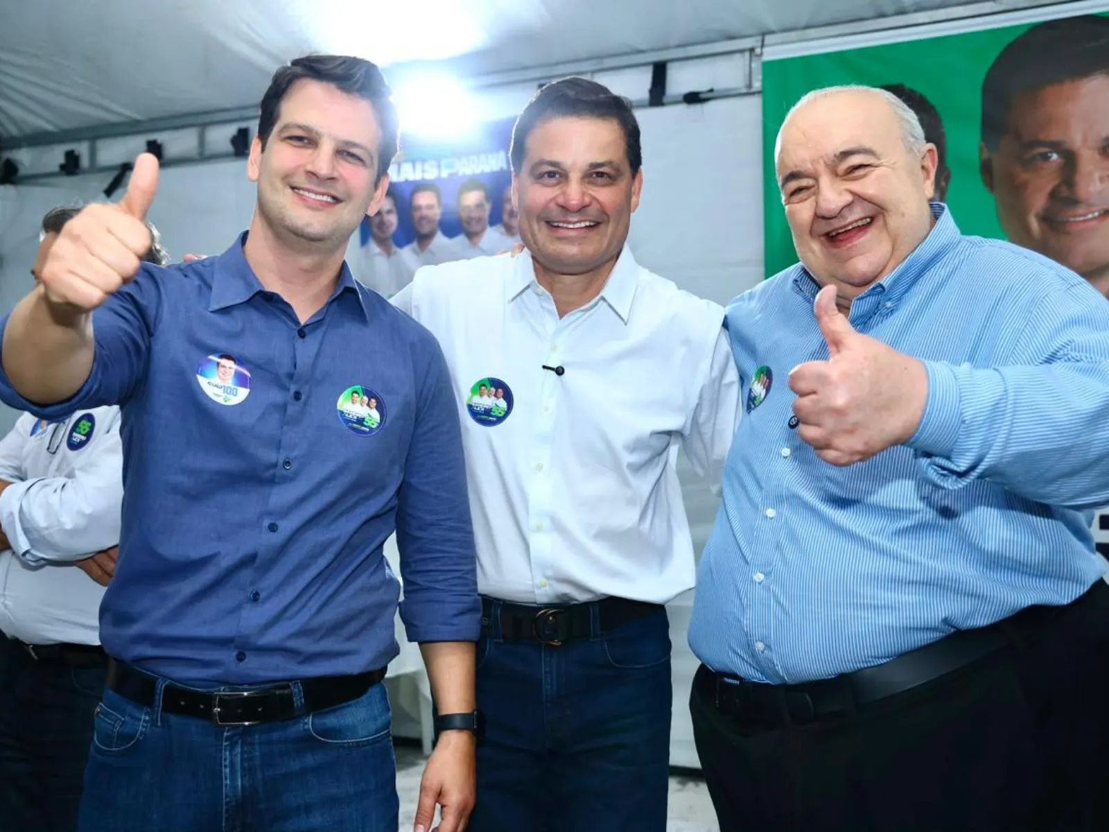 Fotografia mostra três homens posando juntos em um espaço coberto, diante de um painel com imagens de campanha política. Os três sorriem para a câmera. O homem à esquerda usa camisa azul-escura e faz sinal de positivo com a mão direita. O homem ao centro veste camisa branca e está com os braços ao redor dos outros dois. O homem à direita usa camisa listrada azul-clara e também faz sinal de positivo. Os três usam adesivos de campanha nas camisas. Ao fundo, há outras pessoas e materiais de divulgação eleitoral.