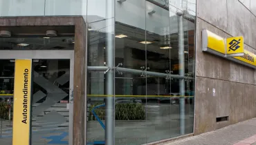 Fotografia mostra a fachada de uma agência do Banco do Brasil, em um prédio com grandes painéis de vidro e paredes de concreto. À direita, há uma placa amarela com o logotipo azul do Banco do Brasil e parte do nome da instituição. Na entrada, um painel vertical amarelo traz a palavra “Autoatendimento”. Através dos vidros, é possível ver a área interna da agência, com caixas de autoatendimento, iluminação e plantas. A calçada é revestida com blocos de concreto e possui uma faixa tátil vermelha próxima ao meio-fio.