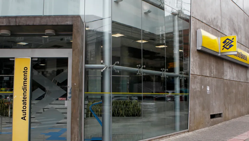 Fotografia mostra a fachada de uma agência do Banco do Brasil, em um prédio com grandes painéis de vidro e paredes de concreto. À direita, há uma placa amarela com o logotipo azul do Banco do Brasil e parte do nome da instituição. Na entrada, um painel vertical amarelo traz a palavra “Autoatendimento”. Através dos vidros, é possível ver a área interna da agência, com caixas de autoatendimento, iluminação e plantas. A calçada é revestida com blocos de concreto e possui uma faixa tátil vermelha próxima ao meio-fio.
