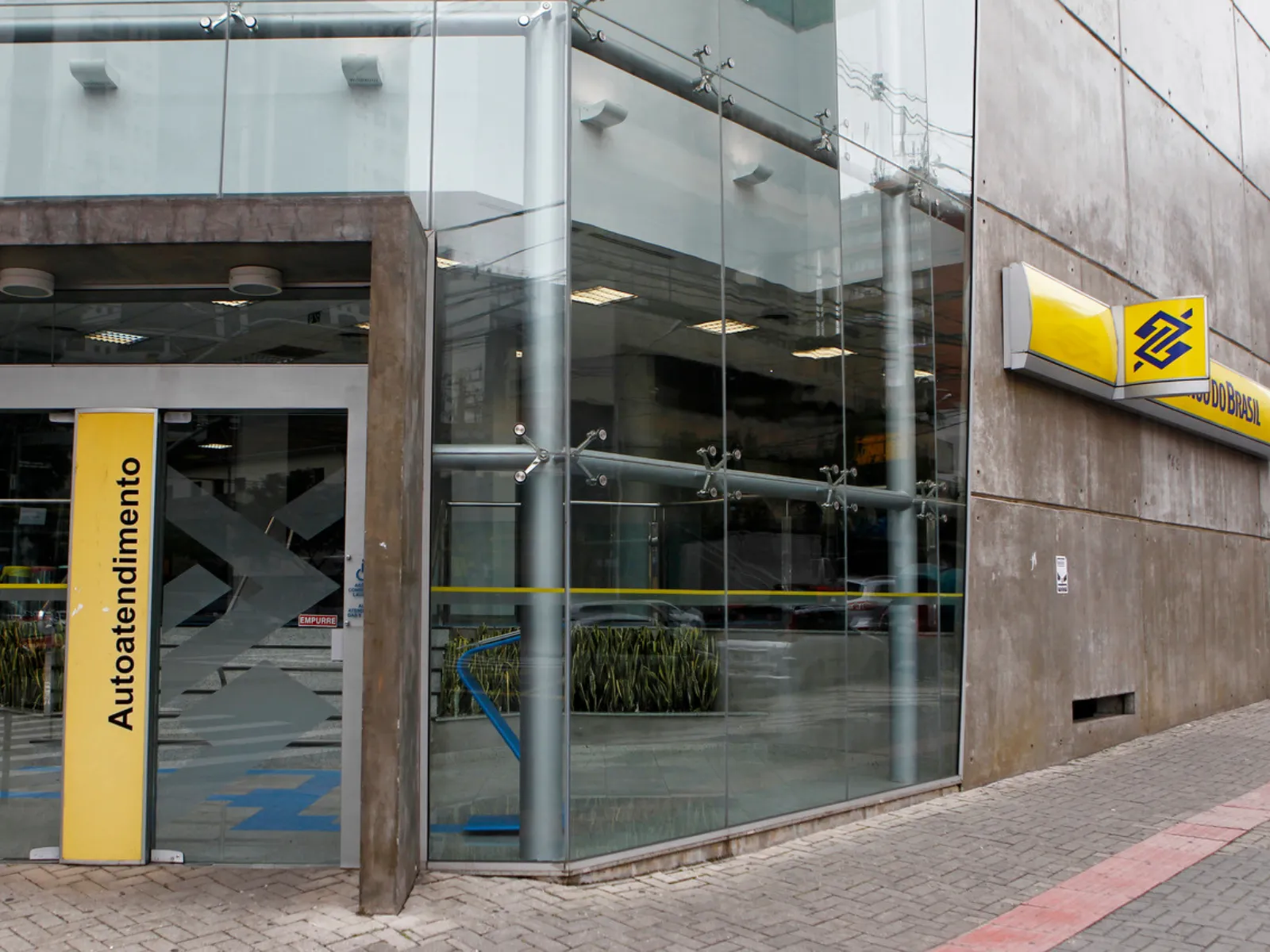 Fotografia mostra a fachada de uma agência do Banco do Brasil, em um prédio com grandes painéis de vidro e paredes de concreto. À direita, há uma placa amarela com o logotipo azul do Banco do Brasil e parte do nome da instituição. Na entrada, um painel vertical amarelo traz a palavra “Autoatendimento”. Através dos vidros, é possível ver a área interna da agência, com caixas de autoatendimento, iluminação e plantas. A calçada é revestida com blocos de concreto e possui uma faixa tátil vermelha próxima ao meio-fio.