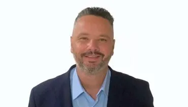 A imagem mostra Luciano Carvalho vestido com camisa azul-claro e paletó preto, em frente a um fundo branco.