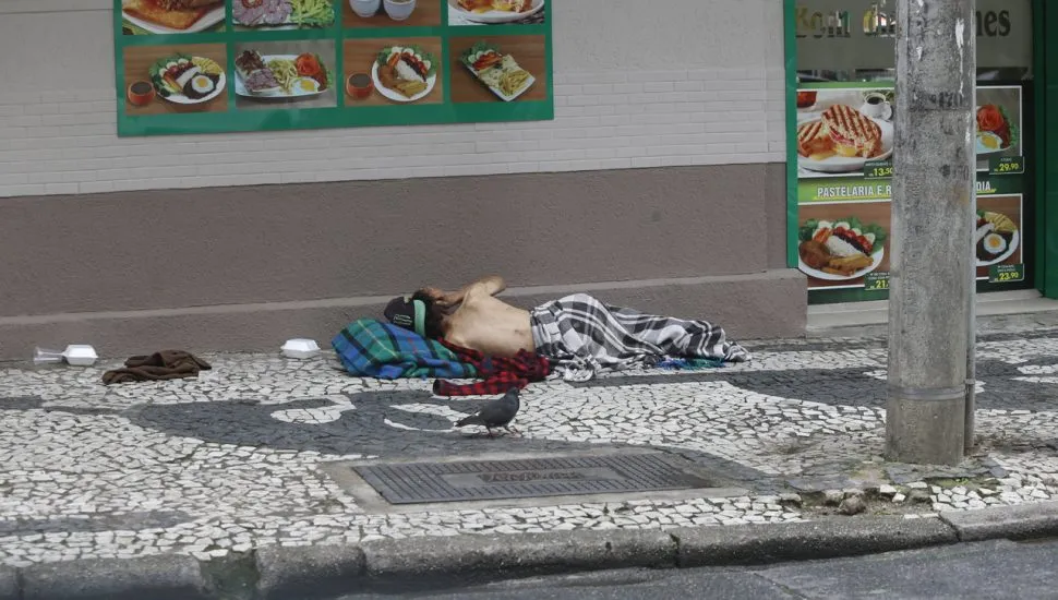 Pessoa em situação de rua dormindo ou descansando na calçada, em frente a um estabelecimento de alimentação, em uma área movimentada de Curitiba.