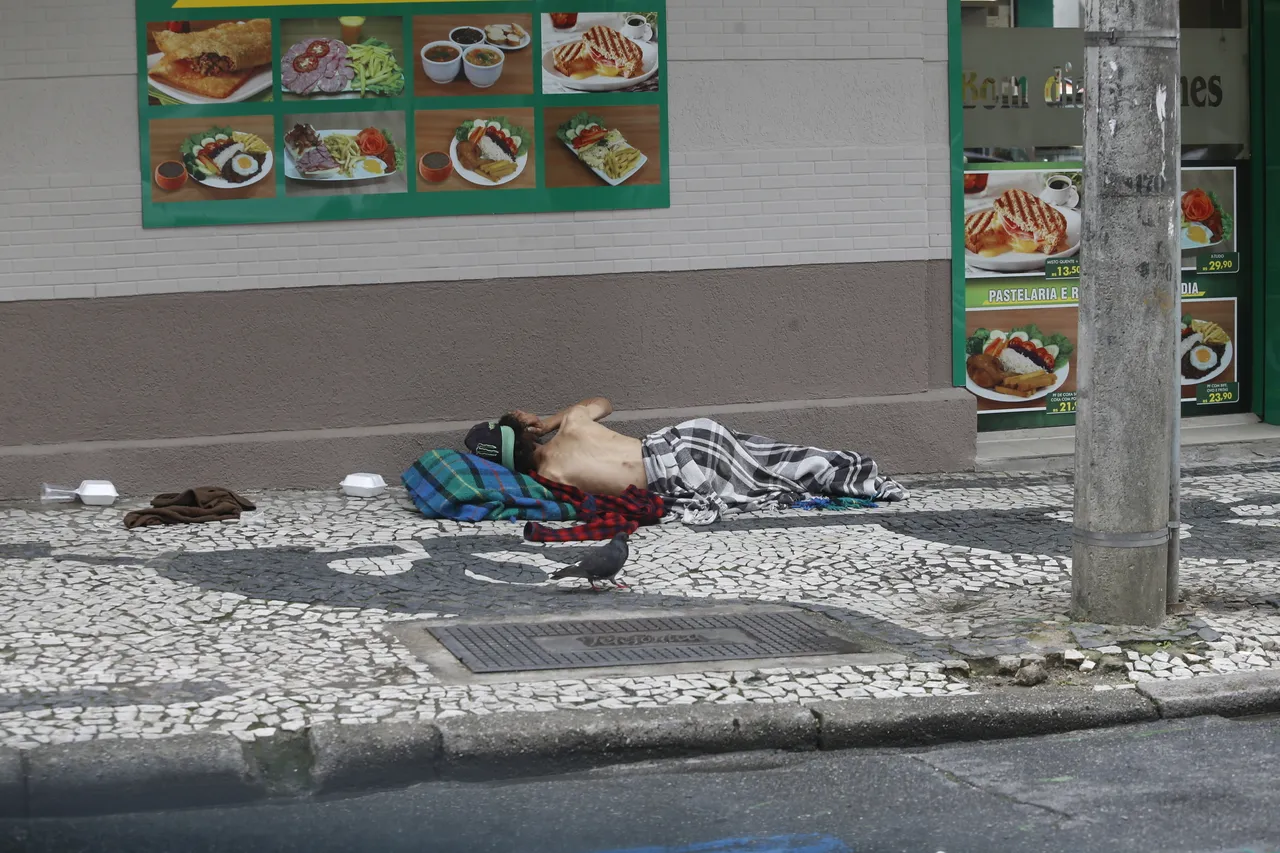 Pessoa em situação de rua dormindo ou descansando na calçada, em frente a um estabelecimento de alimentação, em uma área movimentada de Curitiba.