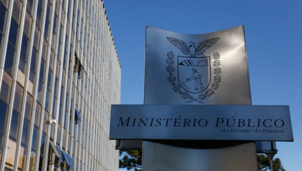 Fotografia mostra a fachada de um prédio institucional alto, com muitas janelas, ao lado de uma placa metálica do Ministério Público do Estado do Paraná. No topo da placa aparece o brasão da instituição, com uma águia sobre um escudo. Abaixo, está escrito “Ministério Público do Estado do Paraná”. Ao fundo, há um céu azul sem nuvens e árvores na parte inferior da imagem.