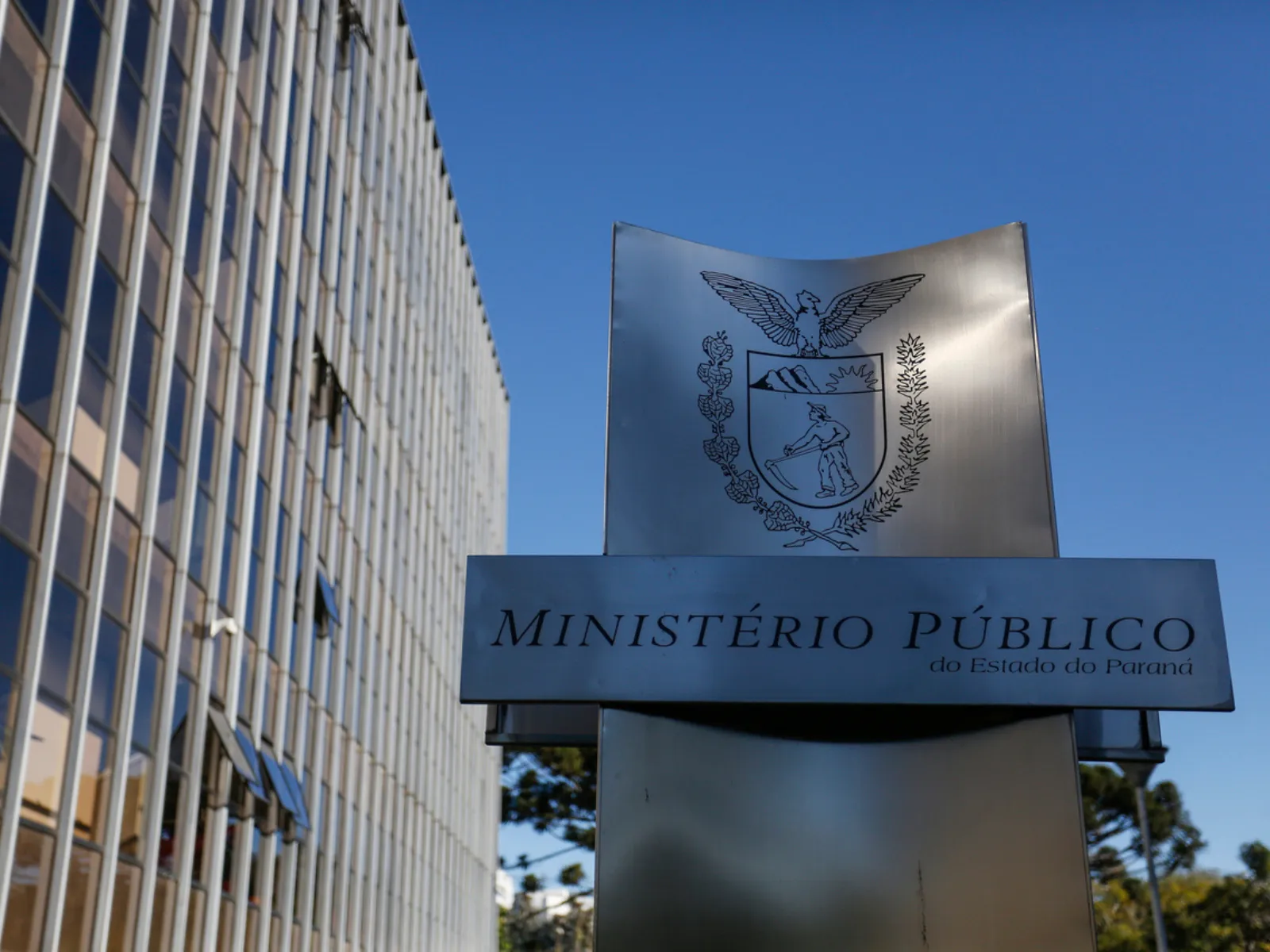 Fotografia mostra a fachada de um prédio institucional alto, com muitas janelas, ao lado de uma placa metálica do Ministério Público do Estado do Paraná. No topo da placa aparece o brasão da instituição, com uma águia sobre um escudo. Abaixo, está escrito “Ministério Público do Estado do Paraná”. Ao fundo, há um céu azul sem nuvens e árvores na parte inferior da imagem.