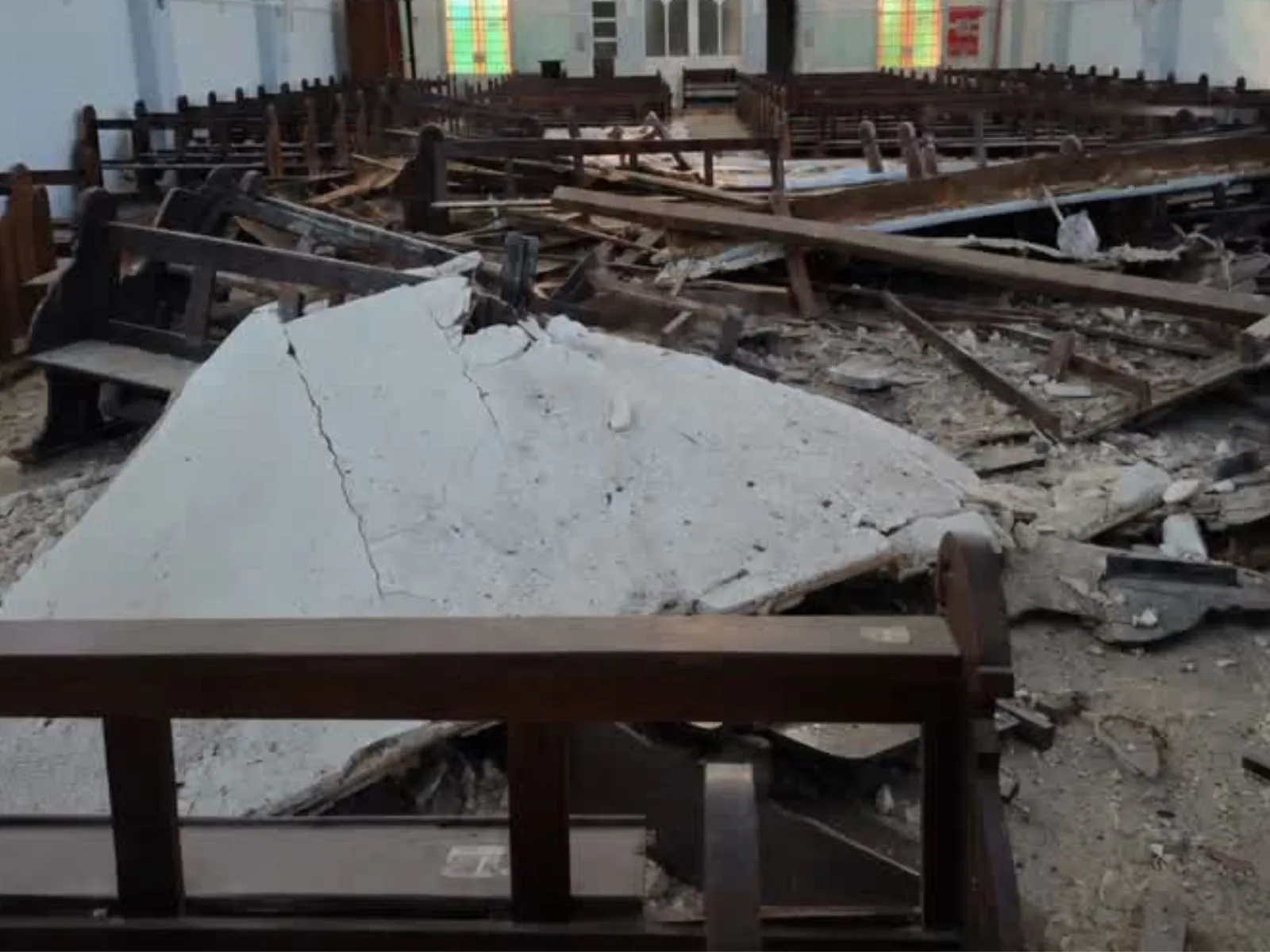 Fotografia do interior de uma igreja após um desabamento no teto. Grandes placas brancas de reboco ou forro caíram sobre os bancos de madeira, quebrando-os e espalhando escombros por boa parte da nave central. Destroços de madeira, entulho e pedaços do forro cobrem o chão entre as fileiras de bancos danificados, alguns tombados e amassados. Ao fundo, veem-se bancos intactos, vitrais coloridos nas janelas laterais e paredes brancas, indicando que a destruição se concentrou em uma área específica do templo.