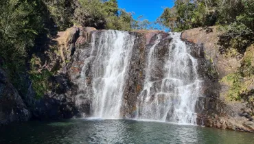Fotografia mostra uma cachoeira em meio à vegetação. A água desce por uma grande parede de rocha escura, formando várias quedas e pequenos cursos que se espalham pela superfície. Na parte inferior, a água se acumula em um poço de tom escuro. Ao lado direito da cachoeira, há vegetação verde e densa. Rochas aparecem em primeiro plano, próximas à água.