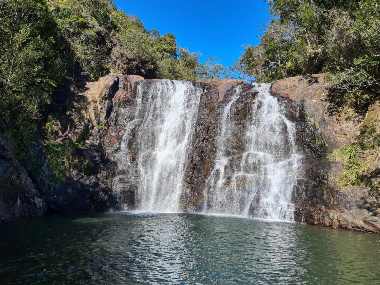 Fotografia mostra uma cachoeira em meio à vegetação. A água desce por uma grande parede de rocha escura, formando várias quedas e pequenos cursos que se espalham pela superfície. Na parte inferior, a água se acumula em um poço de tom escuro. Ao lado direito da cachoeira, há vegetação verde e densa. Rochas aparecem em primeiro plano, próximas à água.