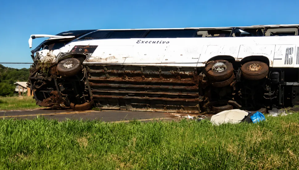 Fotografia mostra um ônibus de viagem branco, identificado como “Executivo”, tombado de lado sobre uma rodovia. O veículo apresenta danos graves na parte dianteira e na lateral, com a estrutura bastante amassada e sujeira de terra e vegetação. As rodas estão voltadas para o lado da câmera. Há pedaços de objetos espalhados próximos ao ônibus. Ao redor, há uma área gramada e, ao fundo, vegetação e algumas construções.