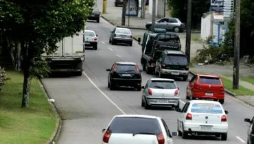 Imagem mostra carros circulando em uma rua de Curitiba, num trecho de curva. Há carros brancos, cinza, pretos e vermelhos, além de um caminhão guincho e árvores.