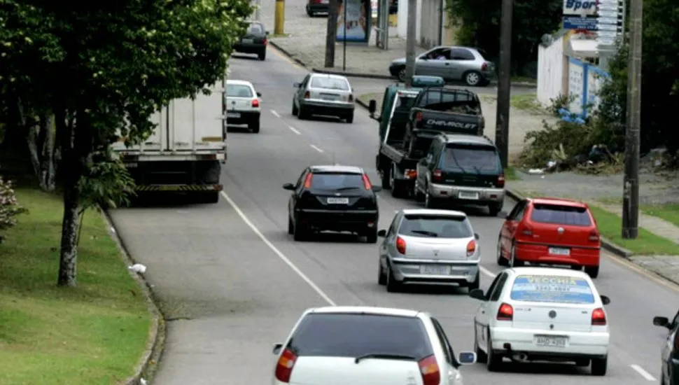 Imagem mostra carros circulando em uma rua de Curitiba, num trecho de curva. Há carros brancos, cinza, pretos e vermelhos, além de um caminhão guincho e árvores.