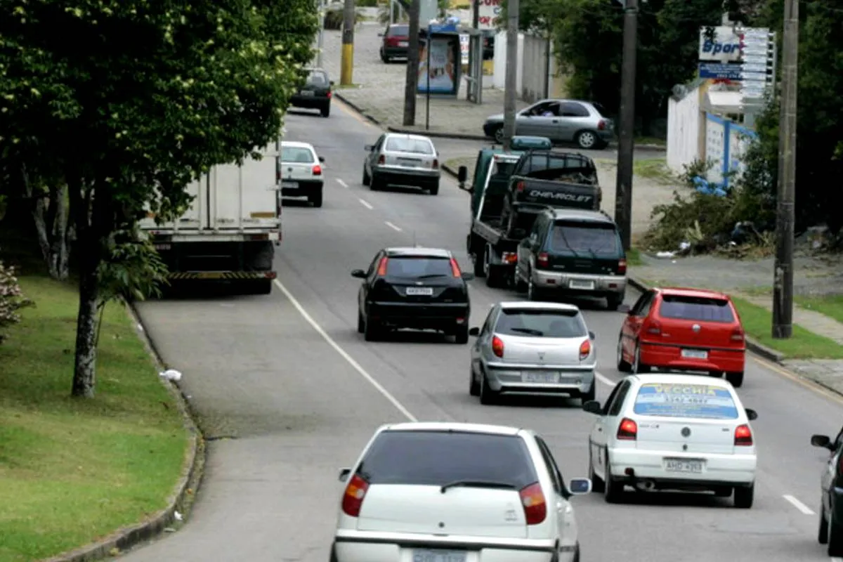 Imagem mostra carros circulando em uma rua de Curitiba, num trecho de curva. Há carros brancos, cinza, pretos e vermelhos, além de um caminhão guincho e árvores.
