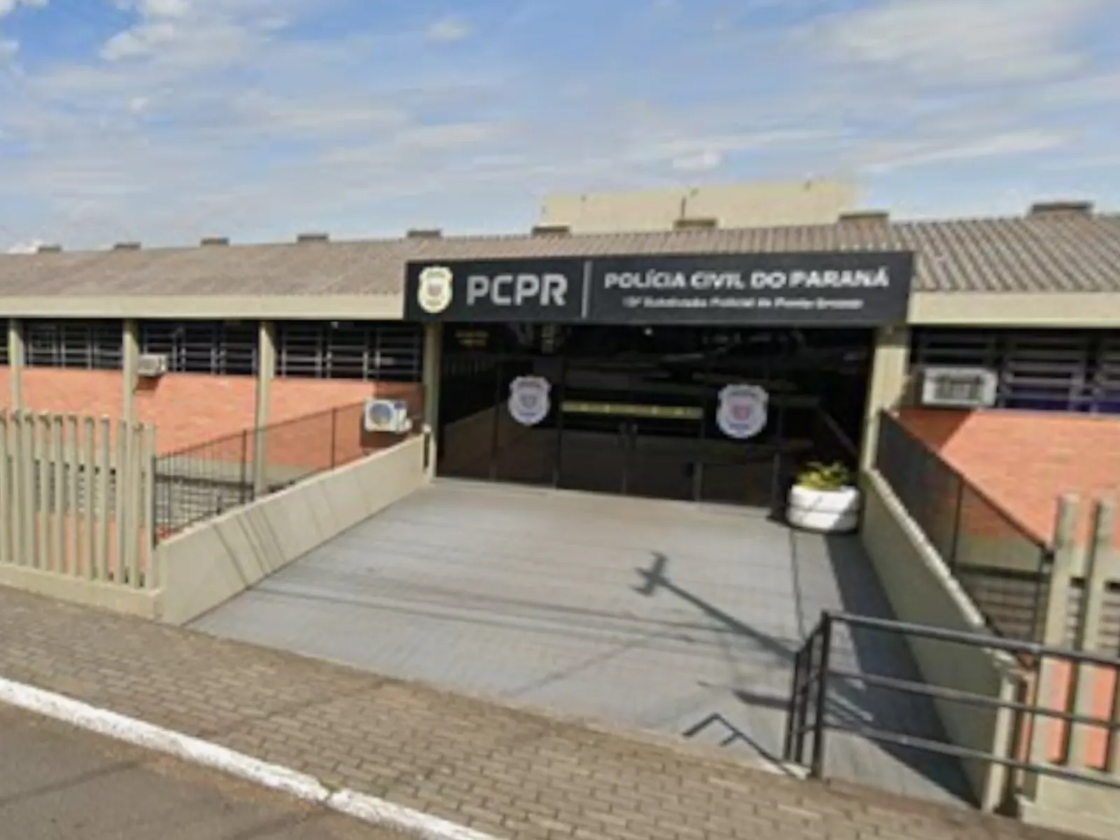 Fotografia mostra a fachada de uma unidade da Polícia Civil do Paraná (PCPR). Na entrada, há uma placa preta com o logotipo da PCPR e a inscrição “Polícia Civil do Paraná”, além de uma identificação da unidade, parcialmente visível. O prédio tem paredes de tijolos aparentes, telhado de telhas e janelas com grades. Uma rampa de acesso leva até a entrada principal, que possui portas de vidro com símbolos da Polícia Civil. Na frente do imóvel há uma calçada e uma rua.