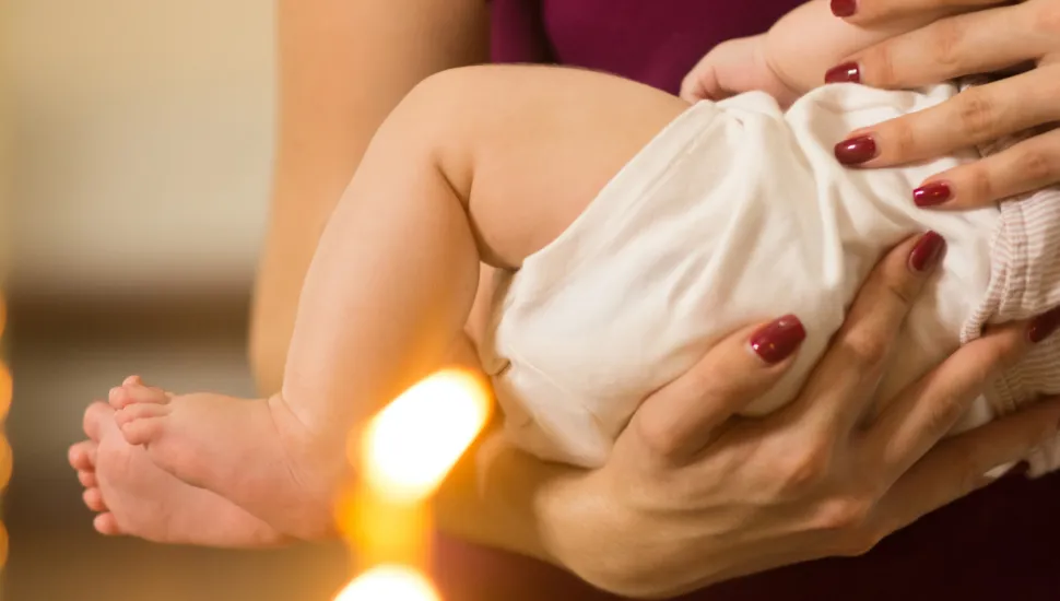 Fotografia em close mostra um bebê sendo segurado no colo por uma pessoa. O bebê veste uma roupa branca e tem as pernas e os pés descalços visíveis. As mãos da pessoa apoiam o corpo da criança; ela usa esmalte vermelho nas unhas. Ao fundo, a pessoa veste uma roupa em tom vinho. A imagem tem iluminação quente e pontos de luz desfocados na parte inferior.