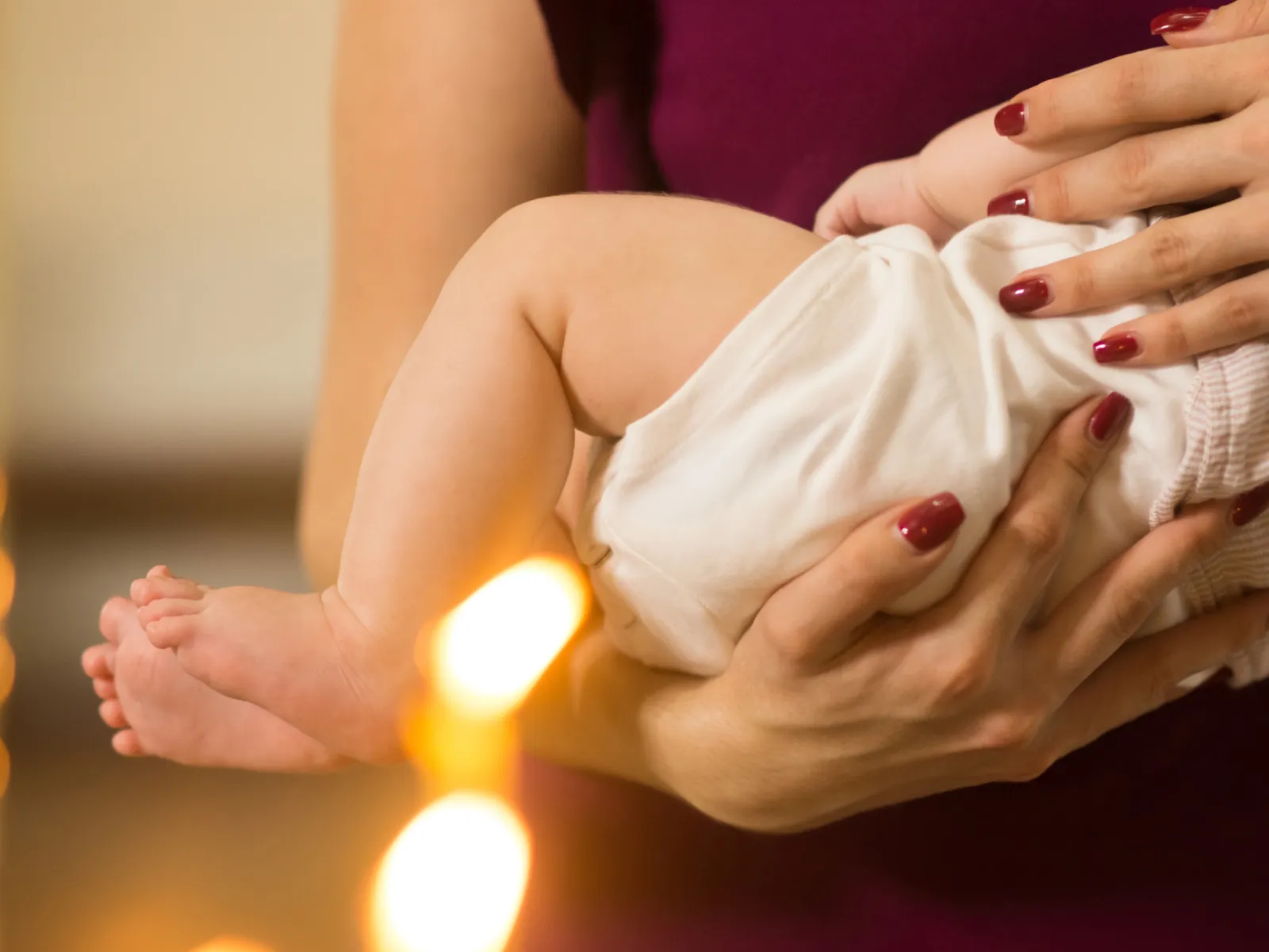 Fotografia em close mostra um bebê sendo segurado no colo por uma pessoa. O bebê veste uma roupa branca e tem as pernas e os pés descalços visíveis. As mãos da pessoa apoiam o corpo da criança; ela usa esmalte vermelho nas unhas. Ao fundo, a pessoa veste uma roupa em tom vinho. A imagem tem iluminação quente e pontos de luz desfocados na parte inferior.