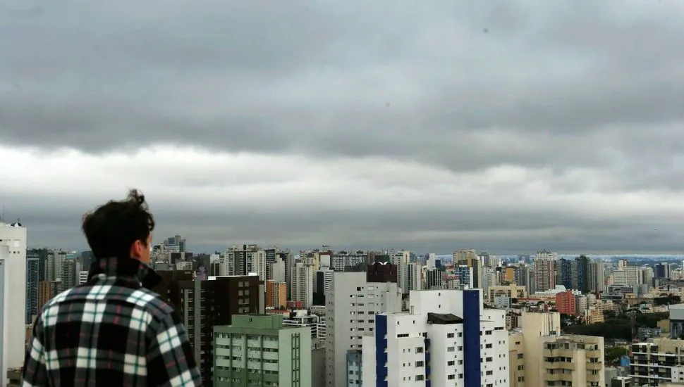 Imagem mostra um homem olhando para o céu carregado de nuvens pesadas e com cara de chuva em cima de uma grande cidade.
