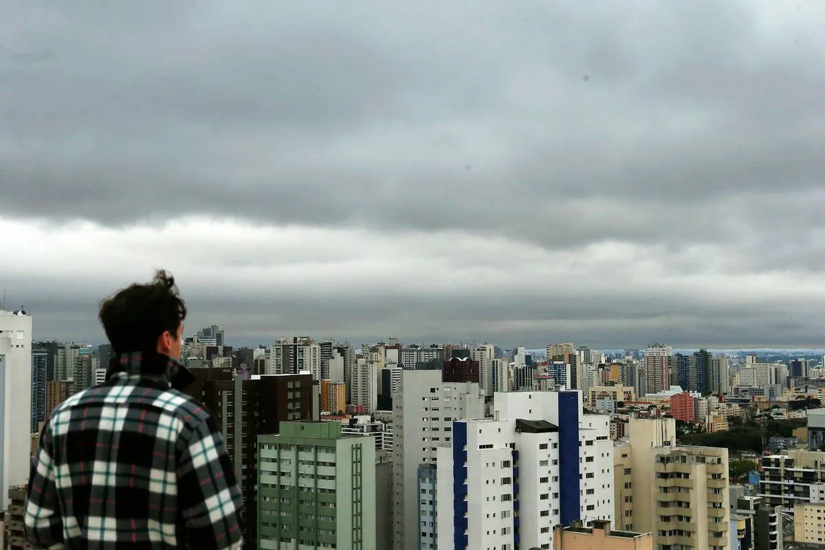 Imagem mostra um homem olhando para o céu carregado de nuvens pesadas e com cara de chuva em cima de uma grande cidade.