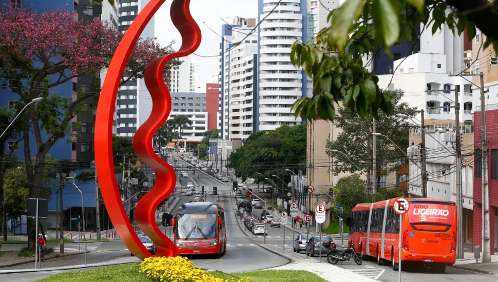 Fotografia urbana diurna de uma avenida movimentada em Curitiba. Em primeiro plano, destaca-se uma grande escultura vermelha curvada em forma de arco sobre um gramado com canteiro de flores amarelas. Ao fundo, veem-se dois ônibus vermelhos do transporte público (Ligeirão), carros em trânsito e edifícios altos sob um céu claro.