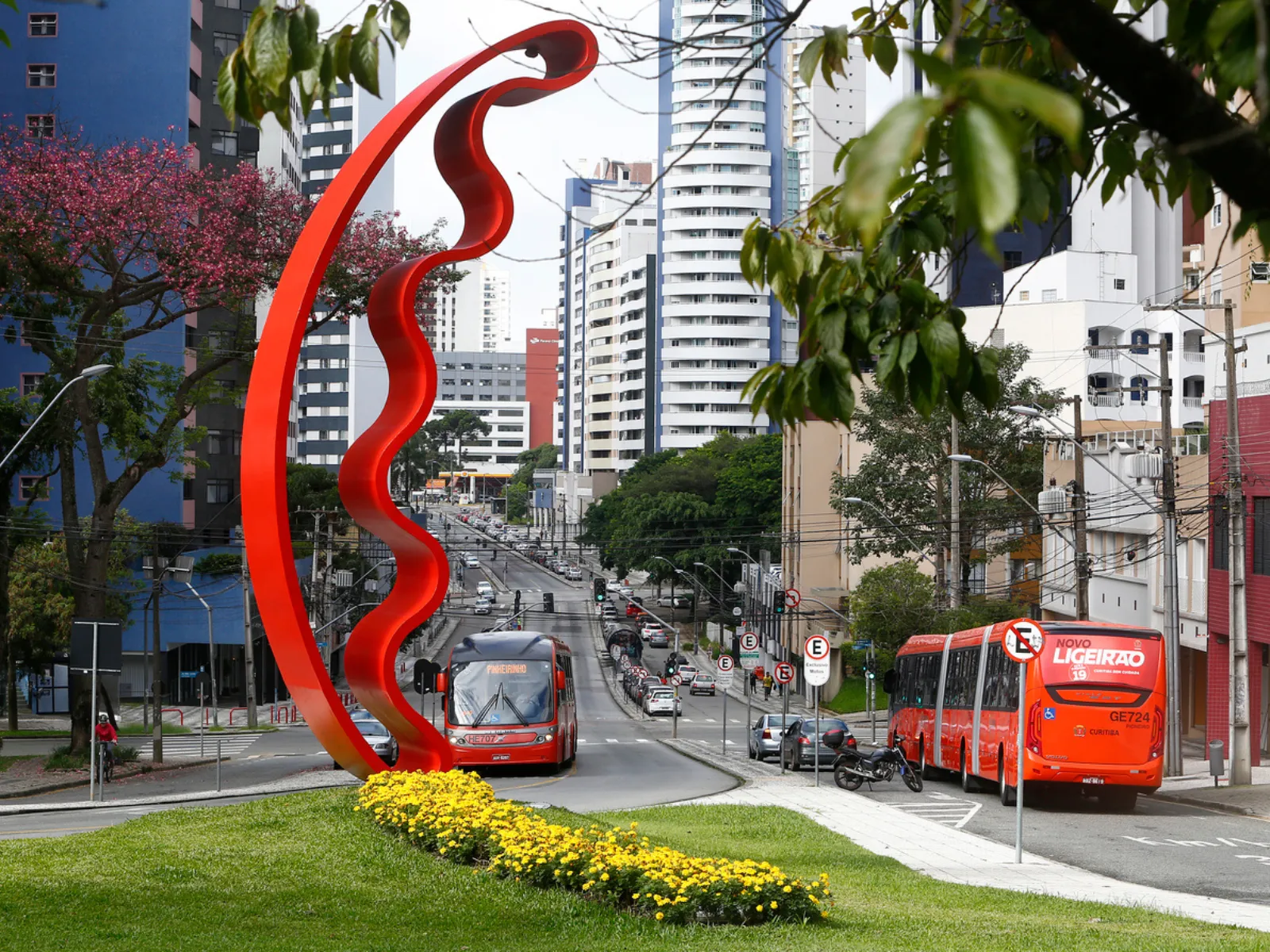 Fotografia urbana diurna de uma avenida movimentada em Curitiba. Em primeiro plano, destaca-se uma grande escultura vermelha curvada em forma de arco sobre um gramado com canteiro de flores amarelas. Ao fundo, veem-se dois ônibus vermelhos do transporte público (Ligeirão), carros em trânsito e edifícios altos sob um céu claro.