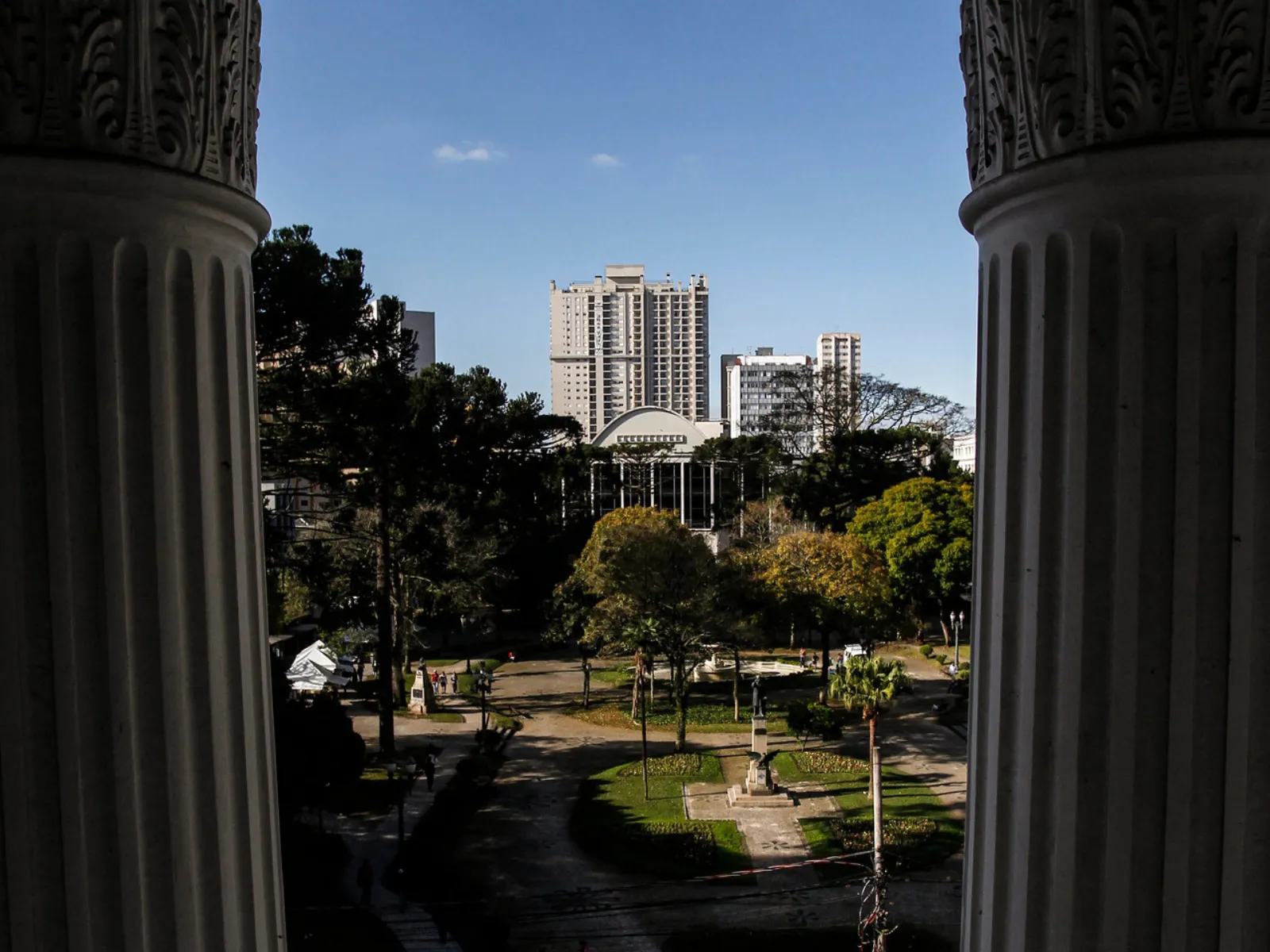 Fotografia de uma praça arborizada vista a partir de um pórtico com colunas clássicas nas laterais da imagem. No centro do parque, há caminhos de pedestres, canteiros de grama, árvores e um monumento. Ao fundo, destacam-se edifícios altos da cidade sob um céu azul e límpido.