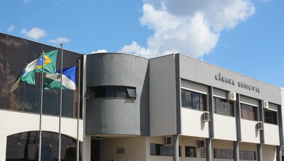 Fotografia em ângulo frontal da fachada de um prédio público moderno de dois andares, com a inscrição "CÂMARA MUNICIPAL" no topo. A estrutura combina paredes brancas, colunas cinza-escuras e grandes janelas espelhadas. À esquerda, em frente ao edifício, há hastes com três bandeiras hasteadas (incluindo a do Brasil e do Paraná) sobre um pequeno gramado. O céu ao fundo é azul com nuvens brancas.