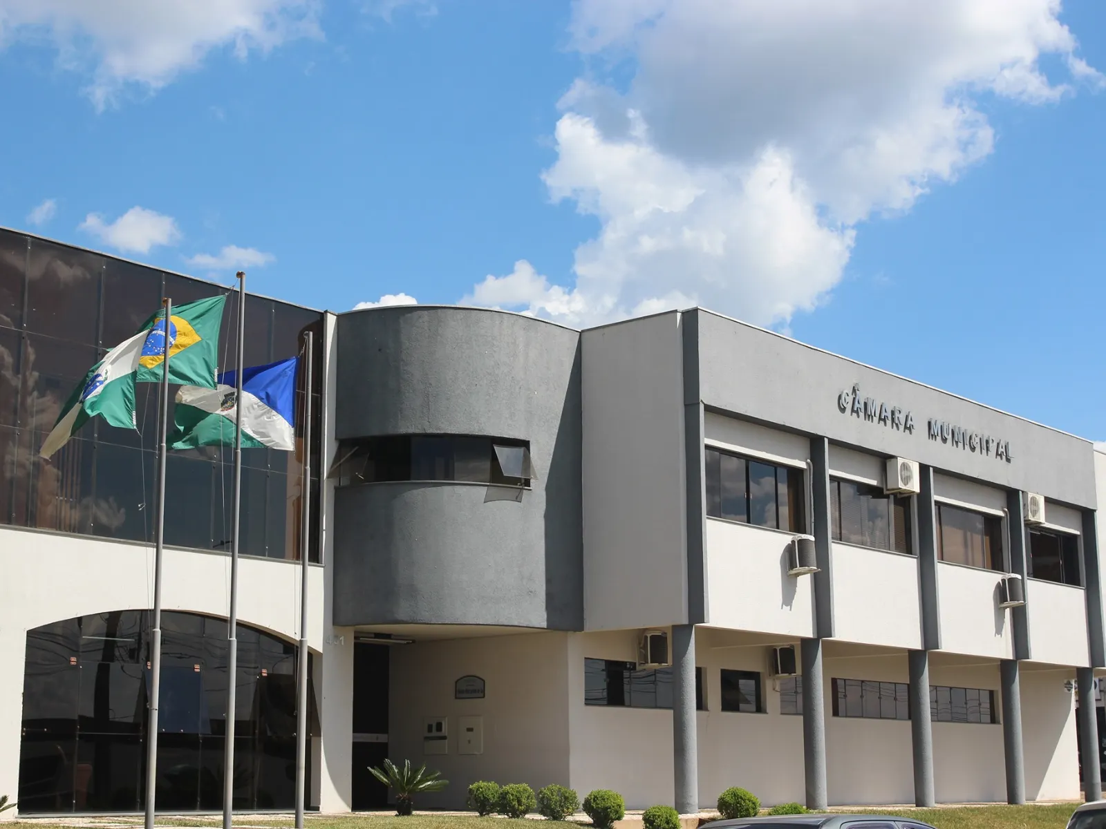 Fotografia em ângulo frontal da fachada de um prédio público moderno de dois andares, com a inscrição "CÂMARA MUNICIPAL" no topo. A estrutura combina paredes brancas, colunas cinza-escuras e grandes janelas espelhadas. À esquerda, em frente ao edifício, há hastes com três bandeiras hasteadas (incluindo a do Brasil e do Paraná) sobre um pequeno gramado. O céu ao fundo é azul com nuvens brancas.