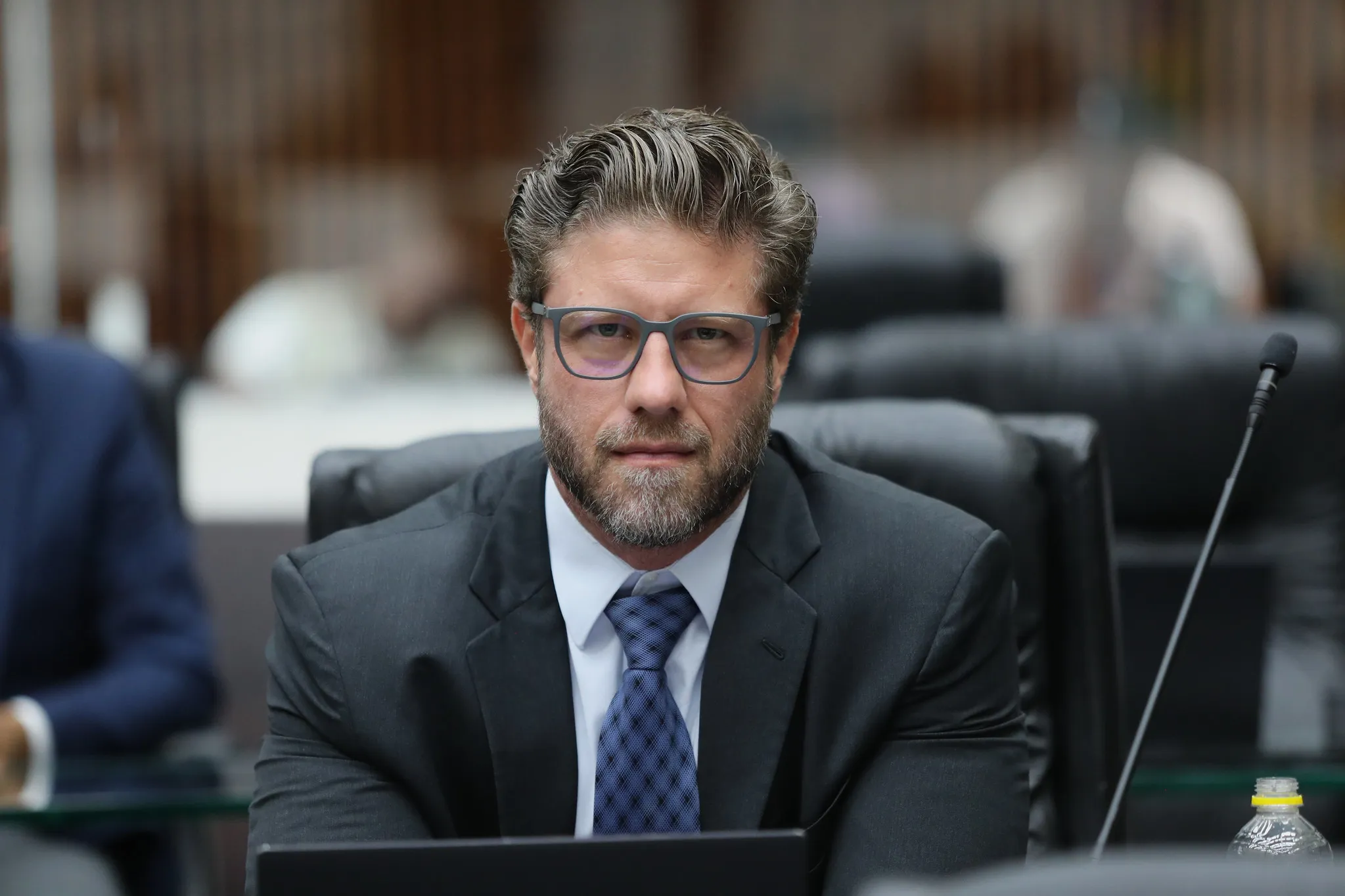 A imagem mostra o deputado estadual Requião Filho sentado e olhando para a câmera. Ele usa uma gravata azul, camisa branca e paletó azul-escuro.