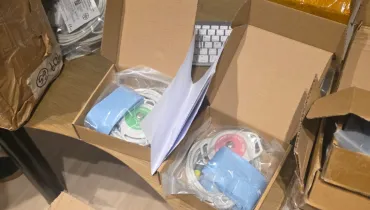 Fotografia em plano superior com foco em duas caixas de papelão abertas sobre uma mesa. Dentro de cada caixa, há equipamentos médicos embalados em sacos plásticos transparentes, contendo cabos, sensores circulares (um com detalhes verdes e outro rosas) e tiras elásticas azuis. Ao redor, há outras caixas de papelão de transporte e sacos plásticos com cabos, com parte de um teclado branco visível ao fundo.