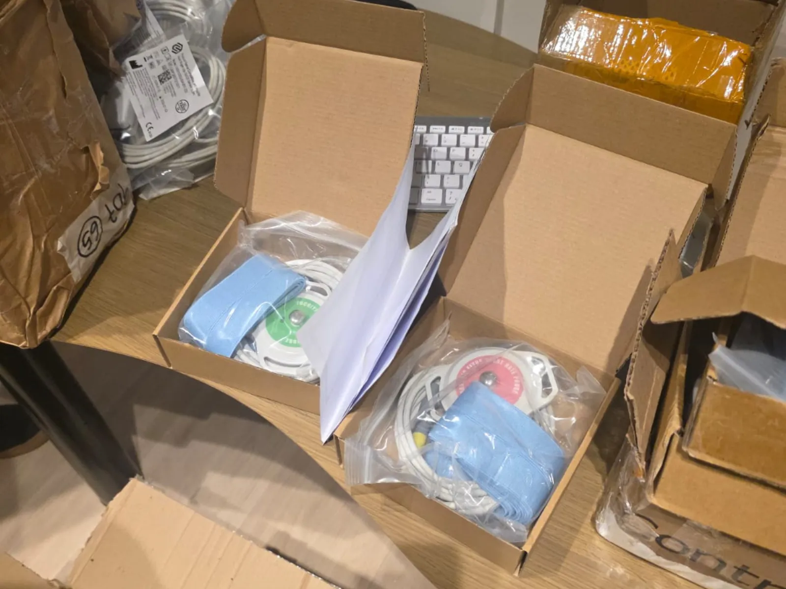 Fotografia em plano superior com foco em duas caixas de papelão abertas sobre uma mesa. Dentro de cada caixa, há equipamentos médicos embalados em sacos plásticos transparentes, contendo cabos, sensores circulares (um com detalhes verdes e outro rosas) e tiras elásticas azuis. Ao redor, há outras caixas de papelão de transporte e sacos plásticos com cabos, com parte de um teclado branco visível ao fundo.