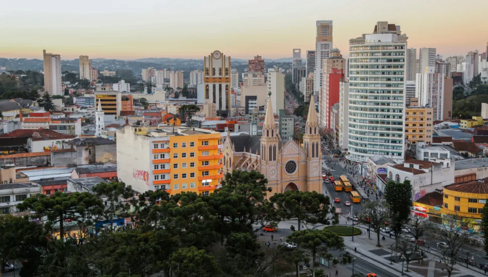 Vista aérea urbana no fim de tarde mostrando o centro de Curitiba. Em destaque no meio da cena, sobressai-se uma catedral em estilo neogótico em tons amarelados, com duas torres pontiagudas. Ao redor e em primeiro plano, há uma praça arborizada com araucárias e calçadas em mosaico. A paisagem é cercada por diversos edifícios residenciais e comerciais de diferentes alturas, sob um céu claro com gradiente alaranjado no horizonte.