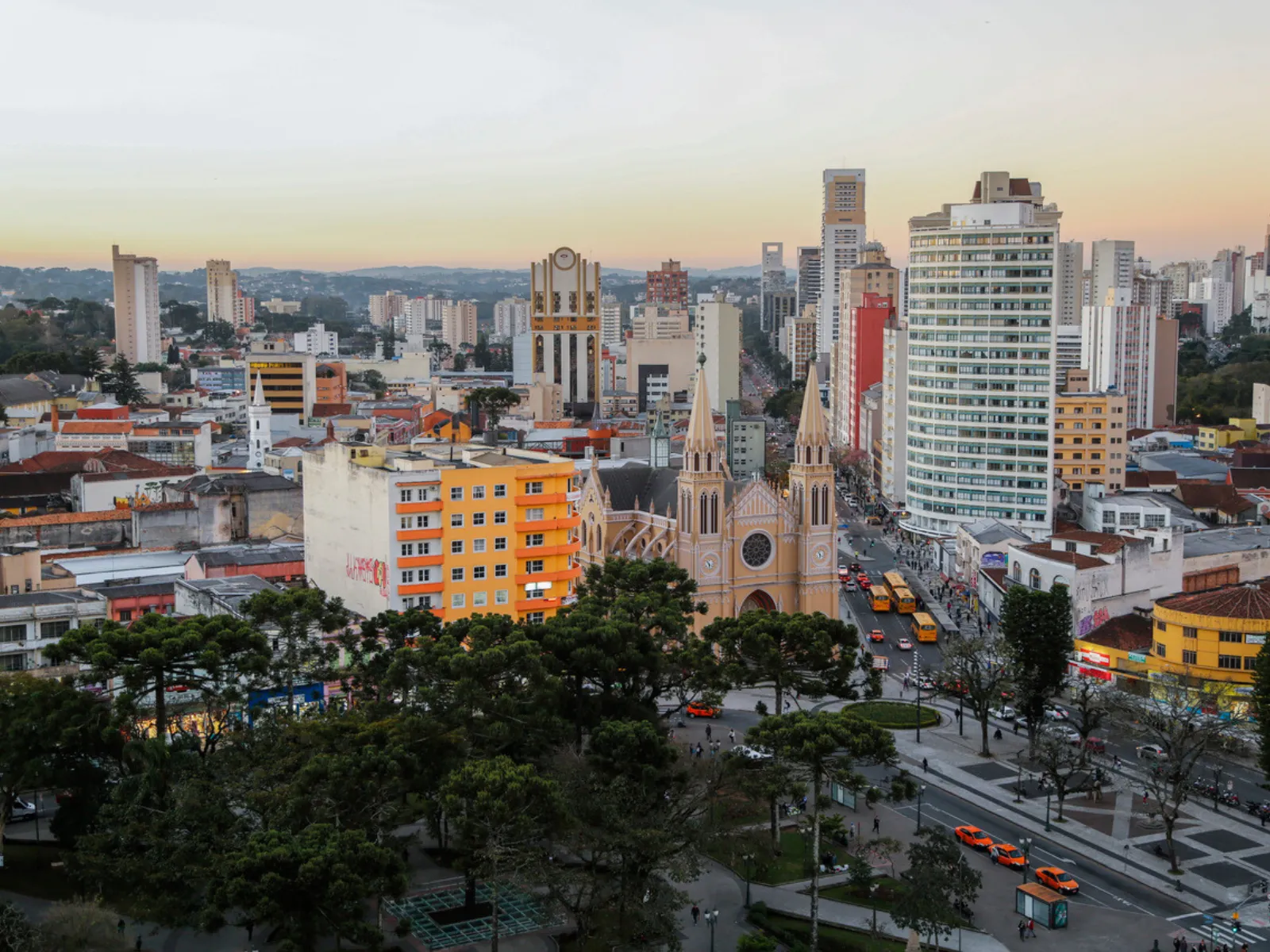 Vista aérea urbana no fim de tarde mostrando o centro de Curitiba. Em destaque no meio da cena, sobressai-se uma catedral em estilo neogótico em tons amarelados, com duas torres pontiagudas. Ao redor e em primeiro plano, há uma praça arborizada com araucárias e calçadas em mosaico. A paisagem é cercada por diversos edifícios residenciais e comerciais de diferentes alturas, sob um céu claro com gradiente alaranjado no horizonte.