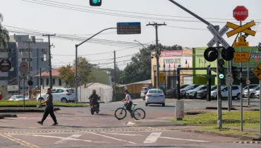 Fotografia de um cruzamento urbano com travessia férrea em dia ensolarado. Em primeiro plano, no lado direito, há uma estrutura de sinalização com semáforo verde, placa de "PARE", cruz de Santo André indicando "VIA CRUZAMENTO FÉRREA" e avisos de trânsito. Na rua, veem-se um pedestre caminhando pela faixa, um motociclista e uma ciclista atravessando o cruzamento. Ao fundo, há carros estacionados e circulando, placas de sinalização viária e imóveis comerciais sob um céu claro.