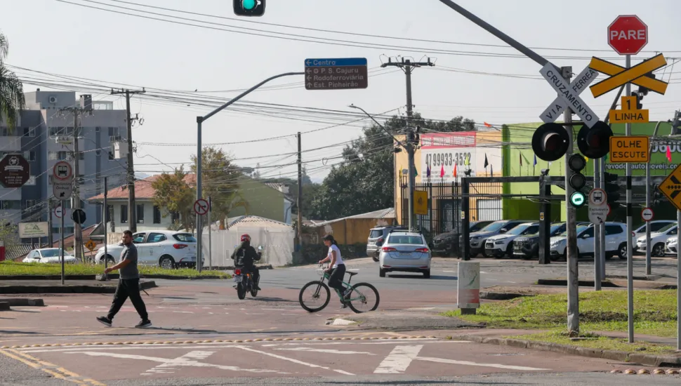 Fotografia de um cruzamento urbano com travessia férrea em dia ensolarado. Em primeiro plano, no lado direito, há uma estrutura de sinalização com semáforo verde, placa de "PARE", cruz de Santo André indicando "VIA CRUZAMENTO FÉRREA" e avisos de trânsito. Na rua, veem-se um pedestre caminhando pela faixa, um motociclista e uma ciclista atravessando o cruzamento. Ao fundo, há carros estacionados e circulando, placas de sinalização viária e imóveis comerciais sob um céu claro.