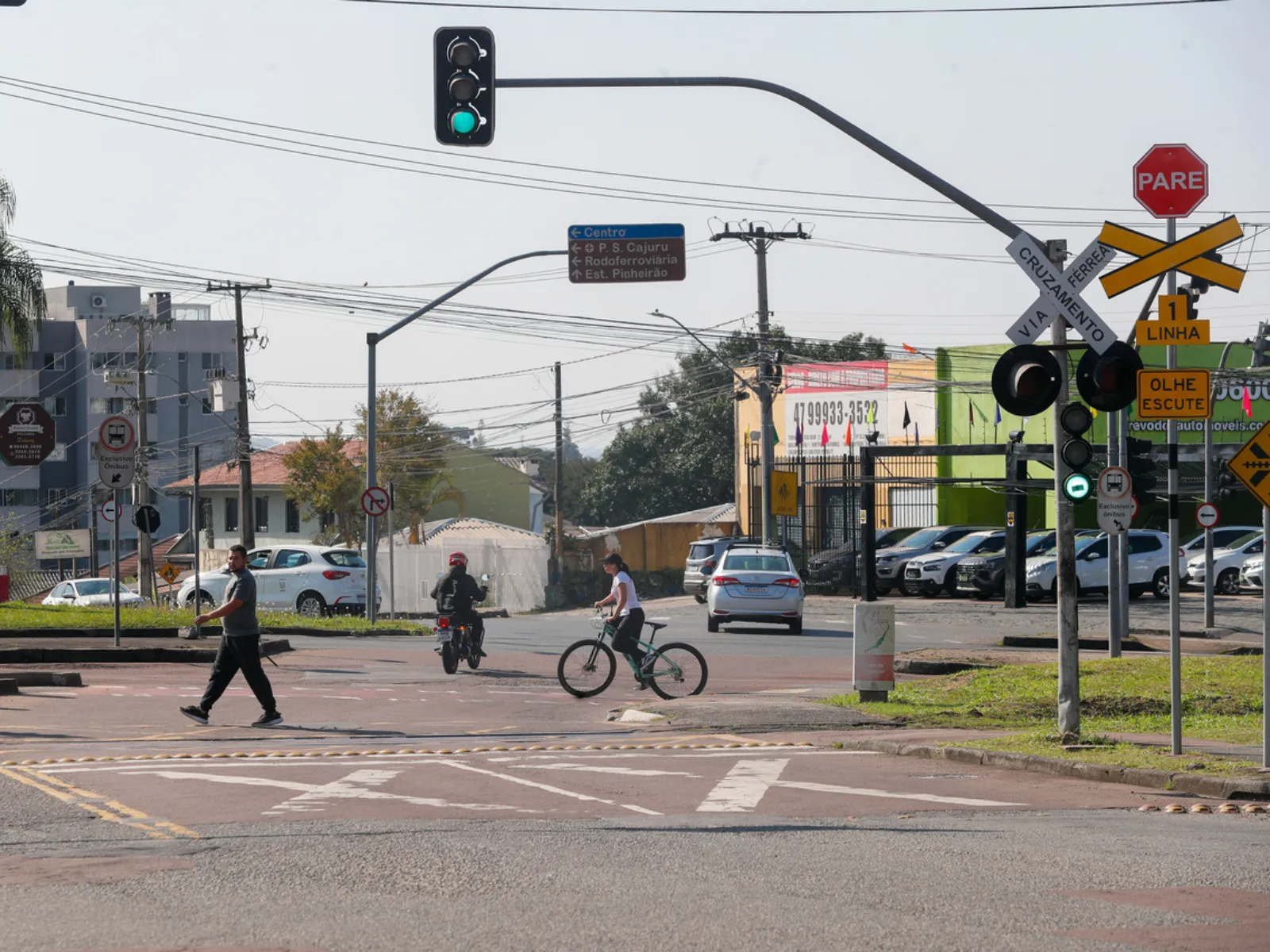 Fotografia de um cruzamento urbano com travessia férrea em dia ensolarado. Em primeiro plano, no lado direito, há uma estrutura de sinalização com semáforo verde, placa de "PARE", cruz de Santo André indicando "VIA CRUZAMENTO FÉRREA" e avisos de trânsito. Na rua, veem-se um pedestre caminhando pela faixa, um motociclista e uma ciclista atravessando o cruzamento. Ao fundo, há carros estacionados e circulando, placas de sinalização viária e imóveis comerciais sob um céu claro.