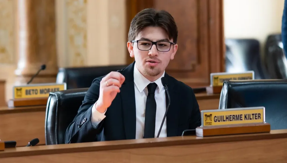 Fotografia em plano médio de um homem jovem de pele clara, cabelos castanho-escuros repartidos ao meio e óculos de grau de armação preta. Ele veste terno preto, camisa social branca e gravata preta, e fala ao microfone enquanto gesticula com a mão direita elevada. Ele está sentado em uma bancada de madeira em um plenário, e à sua frente há uma placa de identificação com o nome "GUILHERME KILTER". Ao fundo, veem-se cadeiras de couro preto, outras bancadas e colunas do ambiente.