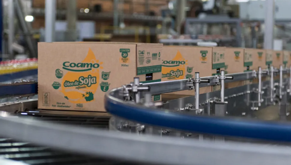 Fotografia em ambiente industrial mostrando uma esteira rolante automatizada transportando várias caixas de papelão. Em primeiro plano e em destaque, há uma caixa da marca Coamo com o texto "Óleo de Soja refinado" impresso em verde e amarelo. As demais caixas seguem em fileira pela linha de produção. Ao fundo, o galpão da fábrica aparece desfocado sob iluminação industrial.
