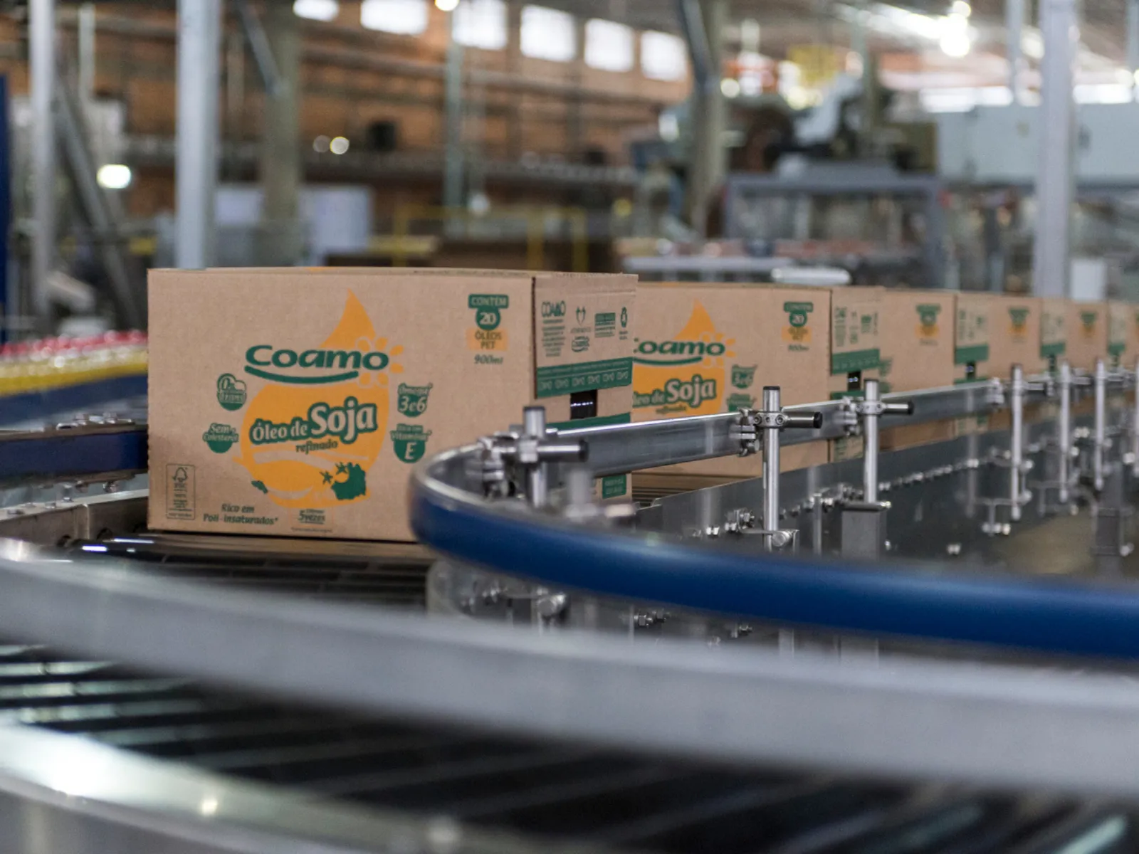 Fotografia em ambiente industrial mostrando uma esteira rolante automatizada transportando várias caixas de papelão. Em primeiro plano e em destaque, há uma caixa da marca Coamo com o texto "Óleo de Soja refinado" impresso em verde e amarelo. As demais caixas seguem em fileira pela linha de produção. Ao fundo, o galpão da fábrica aparece desfocado sob iluminação industrial.