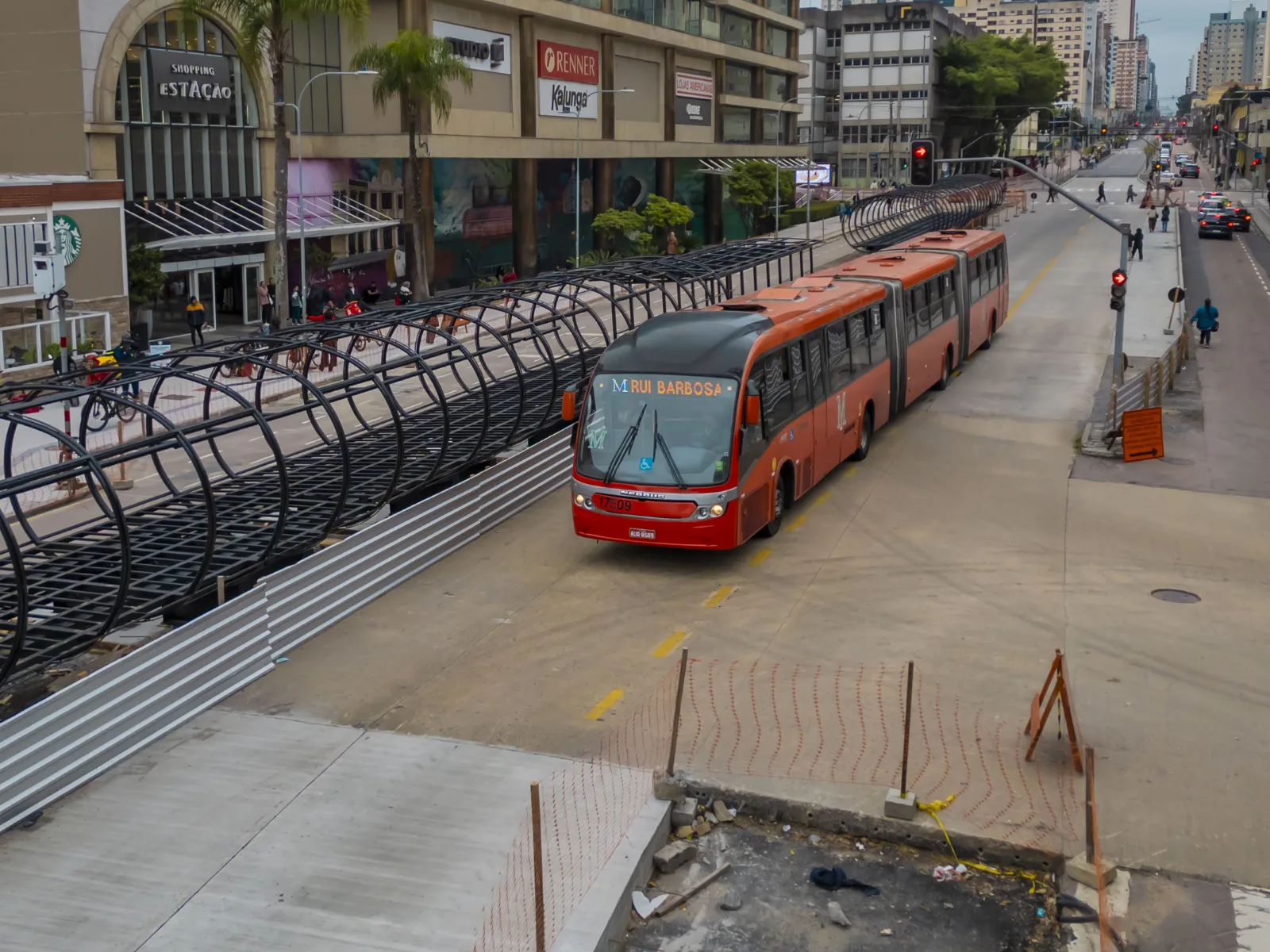 Fotografia em perspectiva elevada de uma canaleta de ônibus em Curitiba. Em destaque no centro, um ônibus biarticulado vermelho trafega pela via de concreto com o letreiro "M RUI BARBOSA". À esquerda do ônibus, há a estrutura metálica cilíndrica de uma estação-tubo em obras de reforma, cercada por tapumes de metal. Ao fundo, no lado esquerdo, vê-se a entrada do Shopping Estação e a fachada de edifícios comerciais sob céu nublado.