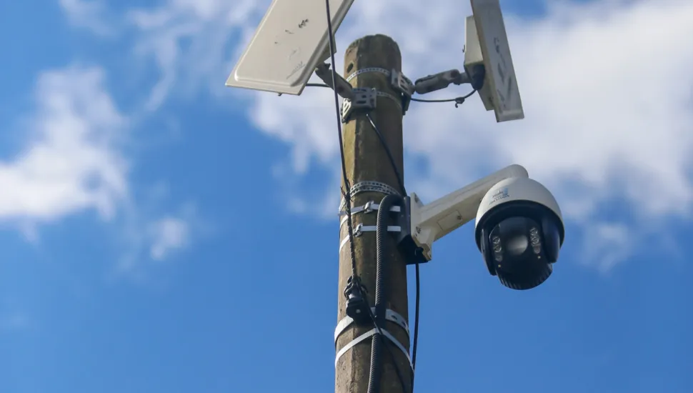 Fotografia em ângulo inferior (vista de baixo para cima) mostrando o topo de um poste de concreto. No poste estão instaladas duas antenas direcionais retangulares brancas na parte superior e uma câmera de monitoramento estilo dome (domo) branca e preta fixada em um suporte lateral logo abaixo. O fundo é composto por um céu azul com nuvens brancas dispersas.