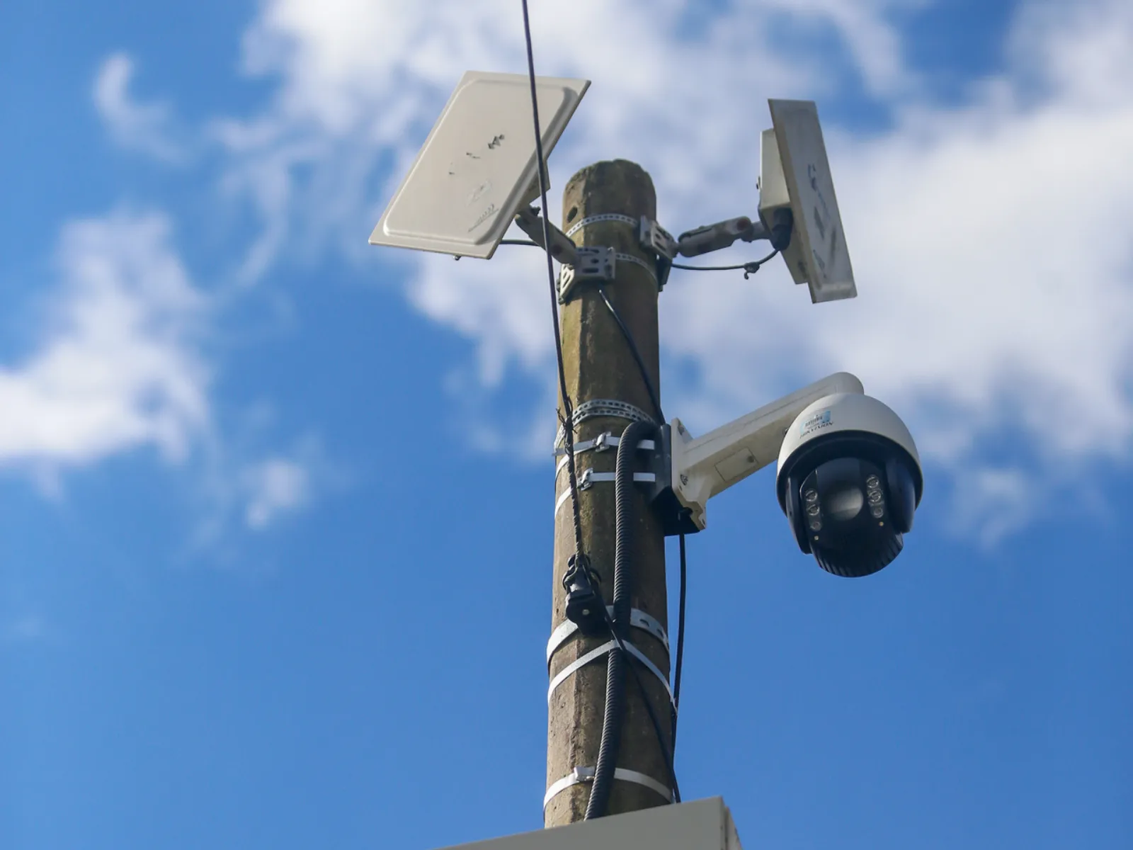 Fotografia em ângulo inferior (vista de baixo para cima) mostrando o topo de um poste de concreto. No poste estão instaladas duas antenas direcionais retangulares brancas na parte superior e uma câmera de monitoramento estilo dome (domo) branca e preta fixada em um suporte lateral logo abaixo. O fundo é composto por um céu azul com nuvens brancas dispersas.