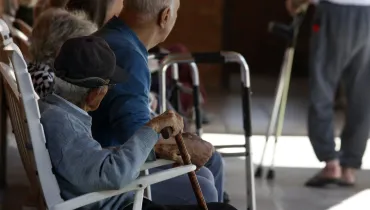Fotografia em ambiente coberto mostrando idosos sentados em fileira em cadeiras brancas, vistos de perfil e de costas. Em primeiro plano, um idoso de boné escuro e casaco cinza segura uma bengala de madeira. Ao seu lado, outro idoso de calvície pronunciada veste um suéter azul. Próximo a eles há um andador metálico e, ao fundo, desfocado, vê-se uma pessoa em pé caminhando com muletas.