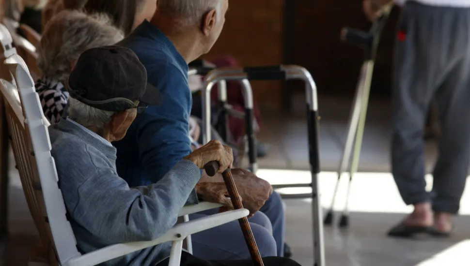 Fotografia em ambiente coberto mostrando idosos sentados em fileira em cadeiras brancas, vistos de perfil e de costas. Em primeiro plano, um idoso de boné escuro e casaco cinza segura uma bengala de madeira. Ao seu lado, outro idoso de calvície pronunciada veste um suéter azul. Próximo a eles há um andador metálico e, ao fundo, desfocado, vê-se uma pessoa em pé caminhando com muletas.