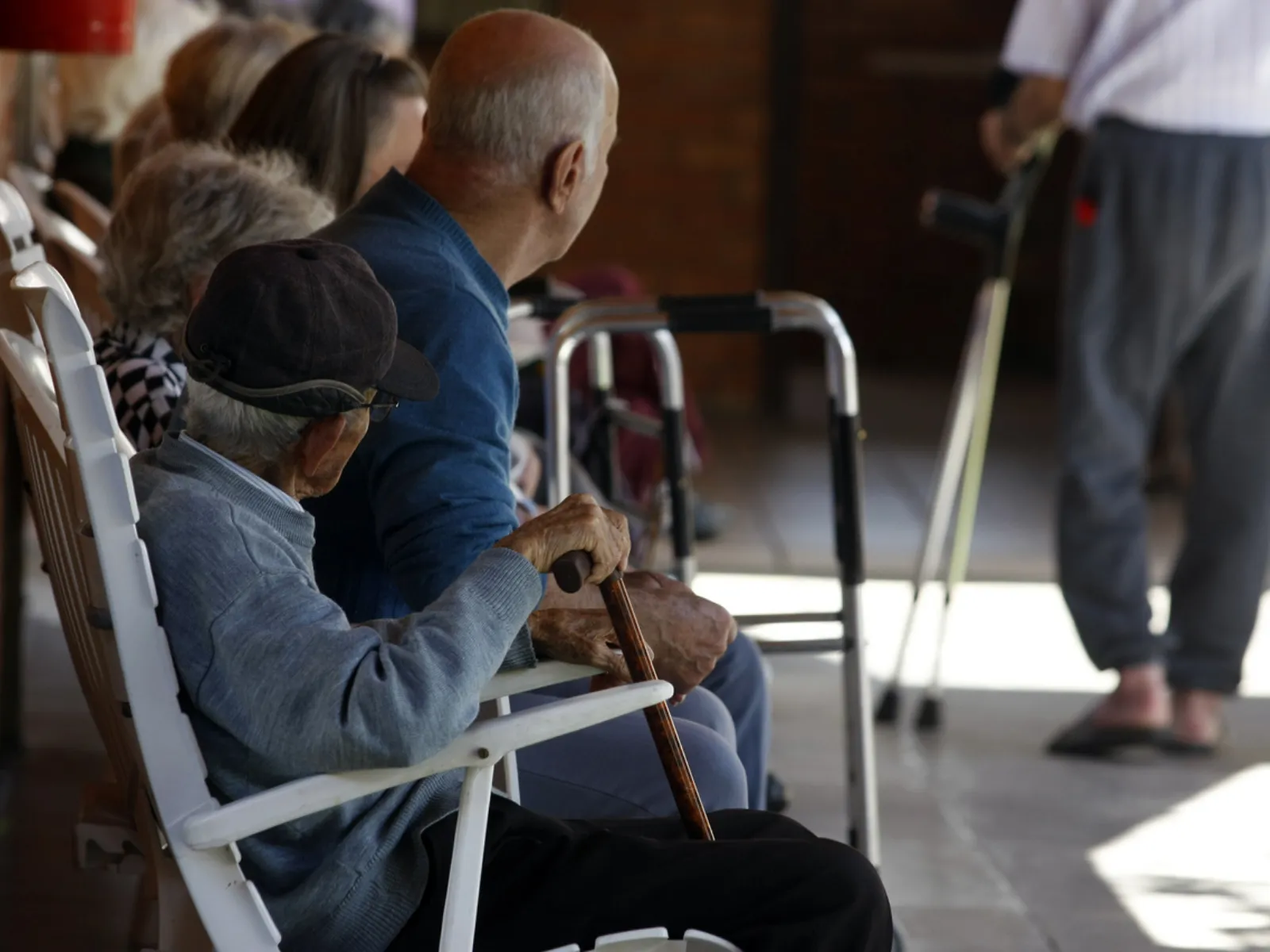 Fotografia em ambiente coberto mostrando idosos sentados em fileira em cadeiras brancas, vistos de perfil e de costas. Em primeiro plano, um idoso de boné escuro e casaco cinza segura uma bengala de madeira. Ao seu lado, outro idoso de calvície pronunciada veste um suéter azul. Próximo a eles há um andador metálico e, ao fundo, desfocado, vê-se uma pessoa em pé caminhando com muletas.