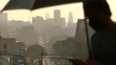Fotografia de uma paisagem urbana vista sob chuva e névoa. Em primeiro plano, no lado direito e em silhueta desfocada, há uma pessoa segurando um guarda-chuva aberto e mexendo no celular. Ao fundo, vê-se uma avenida com trânsito de veículos e faróis acesos sob a chuva pesada, além do contorno de diversos edifícios altos envoltos por uma atmosfera acinzentada e névoa.